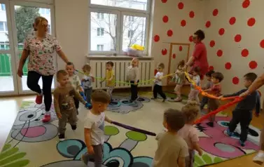 Grupa dzieci wraz z opiekunami stoi na kolorowym dywanie przy wężu spacerowym. 