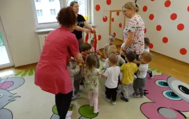 Grupa dzieci wraz z opiekunami stoi na kolorowym dywanie. W ręce trzymają węża spacerowego.