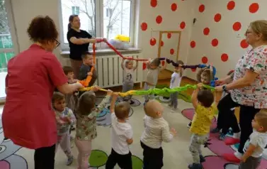Grupa dzieci wraz z opiekunami stoi na kolorowym dywanie. Ich wysoko uniesione ręce trzymają węża spacerowego. 