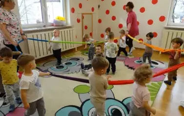 Grupa dzieci wraz z opiekunami stoi na kolorowym dywanie przy wężu spacerowym. 