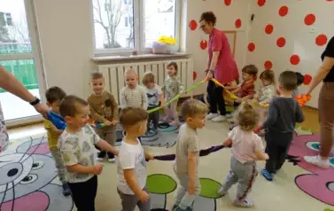 Grupa dzieci wraz z opiekunami stoi na kolorowym dywanie przy wężu spacerowym. 
