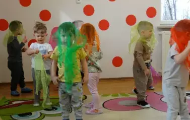 Grupa dzieci stoi na dywanie. Na ich głowach leżą kolorowe chusty.