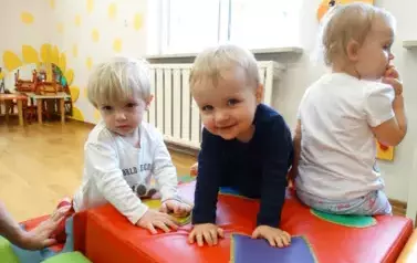 Sala zabaw-na dywanie leżą kolorowe bryły geometryczne, na kt&oacute;rych siedzą dzieci.