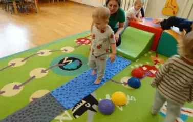 Sala zabaw-na dywanie leżą kolorowe bryły geometryczne i niebieski dywanik, po kt&oacute;rym chodzą dzieci.