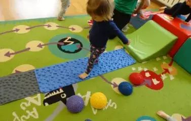 Sala zabaw-na dywanie leżą kolorowe bryły geometryczne i niebieski dywanik, po kt&oacute;rym chodzą dzieci.