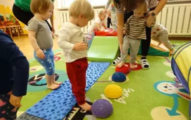 Sala zabaw-na dywanie leżą bryły geometryczne, niebieski dywanik i kolorowe jeżyki, po kt&oacute;rym chodzą dzieci.