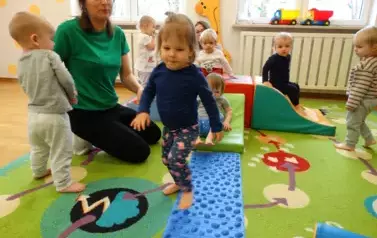Sala zabaw-na dywanie leżą kolorowe bryły geometryczne i niebieski dywanik, po kt&oacute;rym chodzą dzieci.