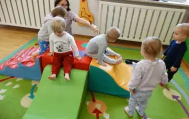 Sala zabaw-na dywanie leżą kolorowe bryły geometryczne a po nich chodzą dzieci.