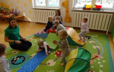 Sala zabaw-na dywanie leżą kolorowe bryły geometryczne i niebieski dywanik, po kt&oacute;rych chodzą dzieci