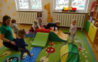 Sala zabaw-na dywanie leżą kolorowe bryły geometryczne i niebieski dywanik, po kt&oacute;rych chodzą dzieci.