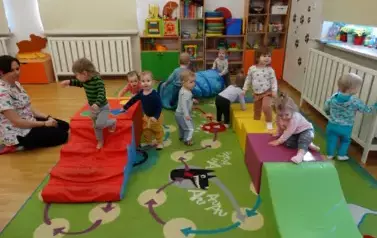 Sala zabaw-na dywanie leżą kolorowe bryły geometryczne a po nich chodzą dzieci.