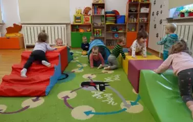 Sala zabaw-na dywanie leżą kolorowe bryły geometryczne a na klęczą siedzą dzieci.