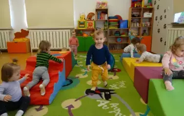 Sala zabaw-na dywanie leżą kolorowe bryły geometryczne a na nich siedzą dzieci.