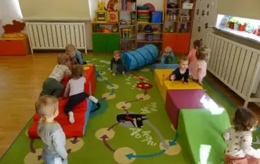 Sala zabaw-na dywanie leżą kolorowe bryły geometryczne a na nich klęczą dzieci.