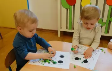 Sala zabaw-dzieci siedzą przy stoliku, na kt&oacute;rym leżą kartki papieru z konturami żaby i zielona bibuła. W ręce trzymają klej.