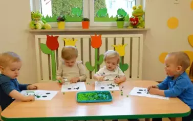 Sala zabaw-grupa dzieci siedzi przy stoliku, na kt&oacute;rym leżą kartki papieru z konturami żaby i zielona bibuła. W ręce trzymają klej.