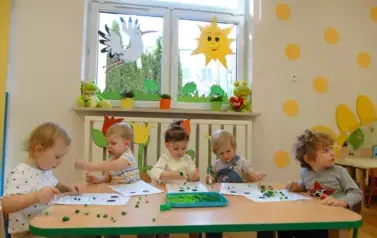 Sala zabaw-dzieci siedzą przy stoliku, na kt&oacute;rym leżą kartki papieru z konturami żaby i zielona bibuła. W ręce trzymają klej.