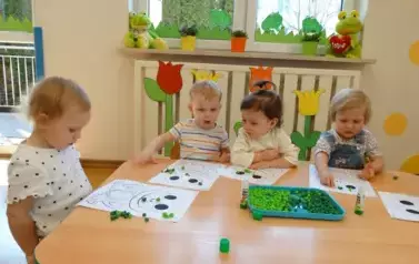 Sala zabaw-dzieci siedzą przy stoliku, na kt&oacute;rym leżą kartki papieru z konturami żaby i zielona bibuła.