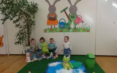 Sala zabaw-dzieci siedzą na krzesełkach, tuż obok nich sztuczny staw z pluszowymi żabami. W tle para kolorowych zajęcy.