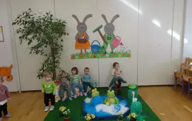 Sala zabaw-dzieci siedzą na krzesełkach, tuż obok nich sztuczny staw z pluszowymi żabami. W tle para kolorowych zajęcy.