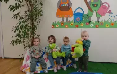 Sala zabaw-dzieci siedzą na krzesełkach, tuż obok nich sztuczny staw z pluszowymi żabami. W tle para kolorowych zajęcy.