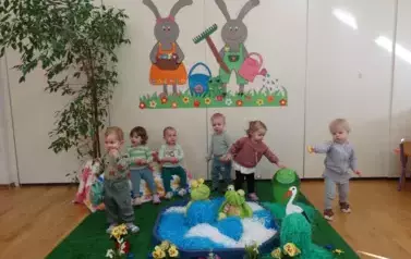 Sala zabaw-dzieci siedzą na krzesełkach, tuż obok nich sztuczny staw z pluszowymi żabami. W tle para kolorowych zajęcy.