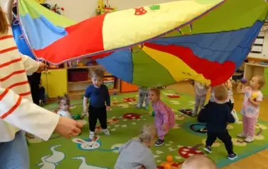 Sala zabaw-opiekunowie unoszą ku g&oacute;rze kolorową chustę animacyjną a pod nią stoi grupka dzieci.