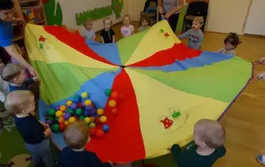 Sala zabaw-opiekunowie i grupka dzieci trzymają w ręce chustę animacyjną, na kt&oacute;rej leżą kolorowe małe piłeczki.