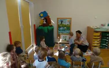 Sala zabaw-opiekun klęczy przy stoliku, na kt&oacute;rym stoi książka. Tuż przy niej chłopiec dotyka reką książki.
