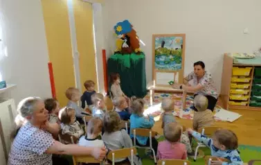 Sala zabaw-opiekun klęczy przy stoliku, na kt&oacute;rym stoi książka. Tuż przy niej na krzesełkach siedzą dzieci.