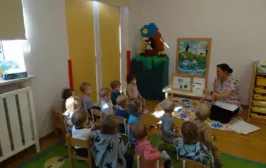 Sala zabaw-opiekun klęczy przy stoliku, na kt&oacute;rym stoi książka. Tuż przy niej na krzesełkach siedzą dzieci.