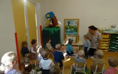 Sala zabaw-opiekun klęczy przy stoliku, na kt&oacute;rym stoi książka. Tuż przy niej stoją dzieci.