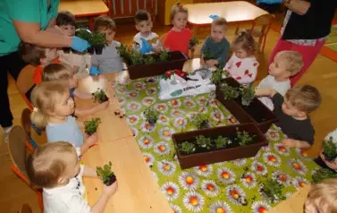 Sala zabaw-dzieci siedzą przy stolikach, na kt&oacute;rych stoją skrzynki z ziemią i kwiatkami. W ręce trzymają sadzonki.