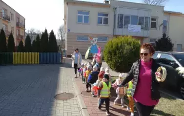 Na pierwszym planie opiekun z tamburynem w ręce. Tuż za nią grupa dzieci trzymająca się węża spacerowego. W tle postać wiosennej Kici Koci. 