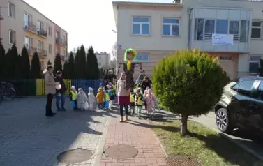 Na pierwszym planie opiekun trzymający w ręce Panią Wiosnę, tuż za nią pozostali opiekunowie i grupa dzieci przy wężu spacerowym.