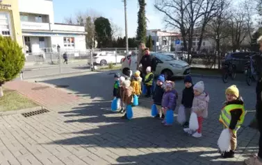 Grupa dzieci stoi przed budynkiem żłobka. W ręce trzymają kolorowe pompony.