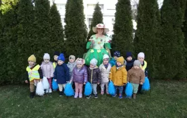 Plac zabaw-grupa dzieci z kolorową Panią Wiosną stoi na zielonej trawie.