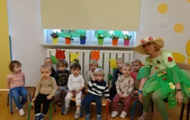 Sala zabaw-Pani Wiosna wraz z grupą dzieci siedzą na krzesełka. W tle kolorowe kwiaty.