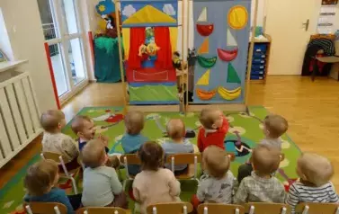 Sala zabaw-grupka dzieci siedzi na krzesełkach. W tle kolorowe kurtyny i pacynki.