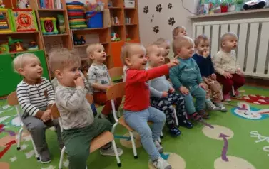 Sala zabaw-grupka uśmiechniętych dzieci siedzi na krzesełkach. W tle kolorowe meble z zabawkami.