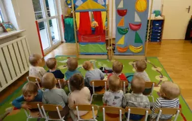 Sala zabaw-grupka dzieci siedzi na krzesełkach. W tle kolorowe kurtyny i pacynki.