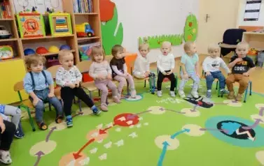 Sala zabaw-grupa dzieci siedzi na krzesełkach. W tle kolorowe meble z zabawkami.