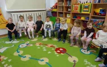 Sala zabaw-grupa dzieci siedzi na krzesełkach. W tle kolorowe meble z zabawkami.