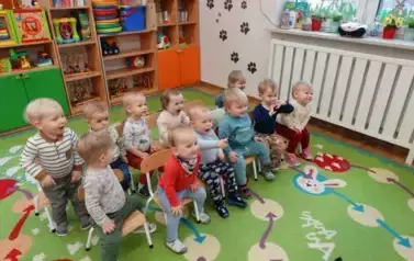 Sala zabaw-grupka uśmiechniętych dzieci siedzi na krzesełkach. W tle kolorowe meble z zabawkami.