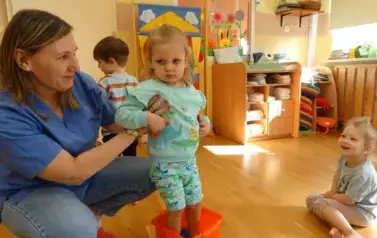 Sala zabaw-opiekun podtrzymuje dziecko, kt&oacute;re bosymi st&oacute;pkami stoi w pomarańczowej misce z niebieską farbą. W tle siedzą dzieci z bosymi st&oacute;pkami.