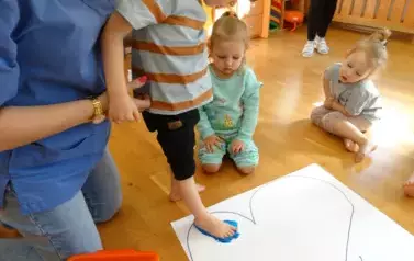 Sala zabaw-opiekun podtrzymuje dziecko, kt&oacute;re bosą niebieską st&oacute;pką stoi na białym papierze. W tle siedzą dzieci z bosymi st&oacute;pkami.