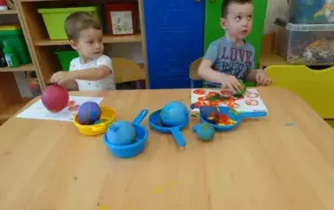 Sala zabaw-dw&oacute;ch chłopc&oacute;w siedzi przy stoliku, na kt&oacute;rym leżą farby. W ręce trzymają małe baloniki i dotykają nimi kartek leżących tuż przd nimi.