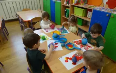 Sala zabaw-grupka dzieci siedzi przy stoliku, na kt&oacute;rym leżą farby. W ręce trzymają małe baloniki i dotykają nimi kartek leżących tuż przd nimi.