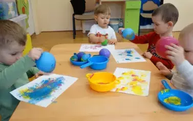 Sala zabaw-grupka dzieci siedzi przy stoliku, na kt&oacute;rym leżą farby. W ręce trzymają małe baloniki i dotykają nimi kartek leżących tuż przd nimi.