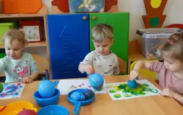 Sala zabaw-tr&oacute;jka dzieci siedzi przy stoliku, na kt&oacute;rym leżą farby. W ręce trzymają małe baloniki i dotykają nimi kartek leżących tuż przd nimi.